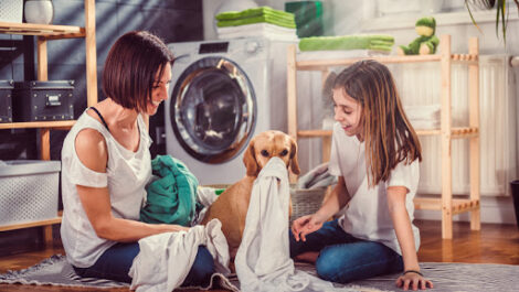 A mother and daughter smiling and laughing as their dog plays with laundry in the laundry room.