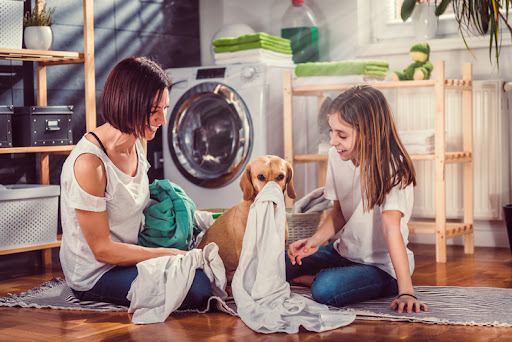 A mother and daughter smiling and laughing as their dog plays with laundry in the laundry room.