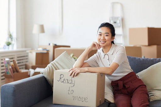 A college student sitting on her couch next to a taped, labeled box for summer storage.