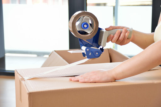 A young woman closing a cardboard box with tape at home.