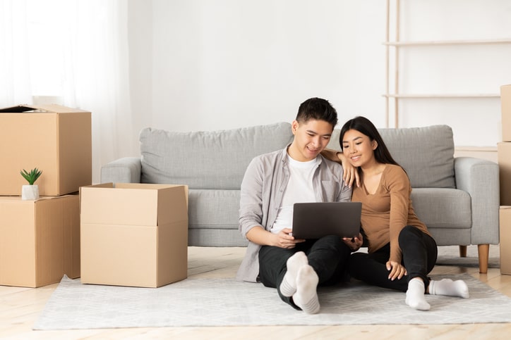 A smiling couple sitting on the floor next to a stack of moving boxes.