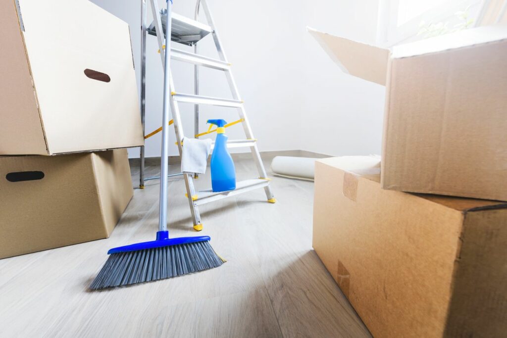 A broom and spray bottle of cleaning solution are propped up on a ladder, surrounded by moving boxes.