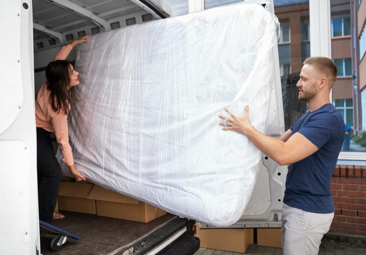 A man and a woman move a mattress wrapped in plastic out of the back of a moving truck.