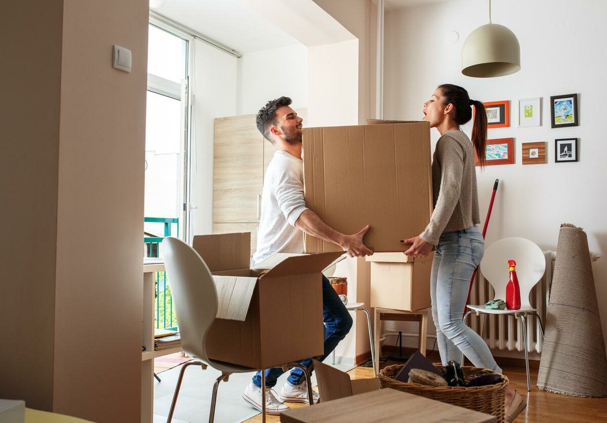 A man and woman carry a large box into the living room of their new apartment, navigating around chairs, furniture, and other boxes.
