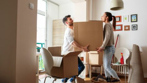 A man and woman carry a large box into the living room of their new apartment, navigating around chairs, furniture, and other boxes.