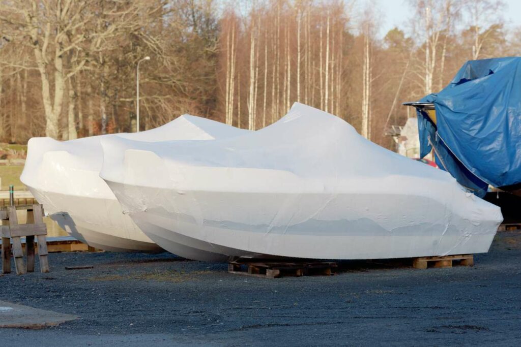 Two boats sit in parking lot and are surrounded by a white boat cover