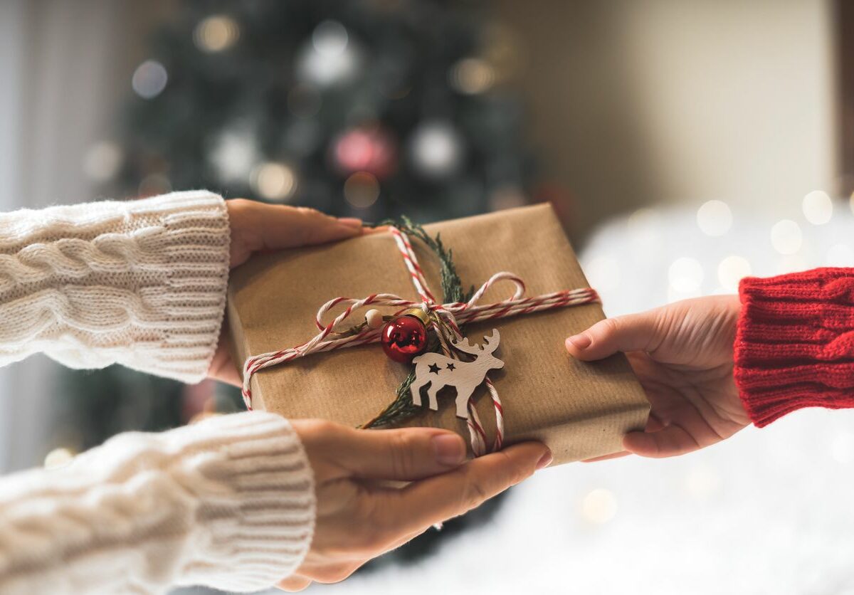 A person hands a festively wrapped gift to a friend while both stand in front of a Christmas tree.