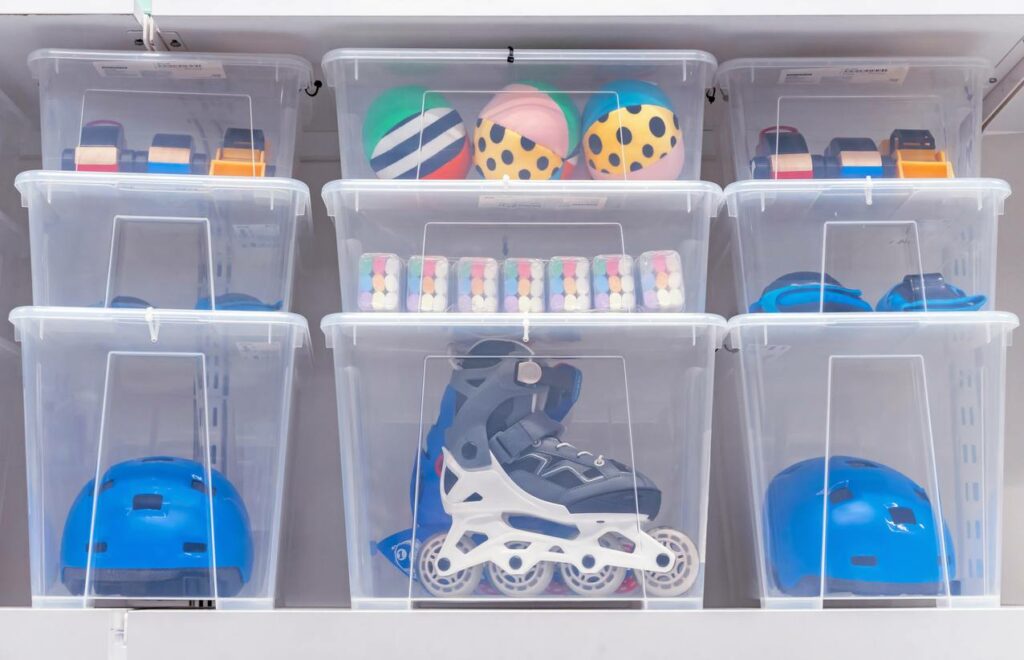 Clear plastic bins hold outdoor equipment including helmets, roller blades, and chalk. 