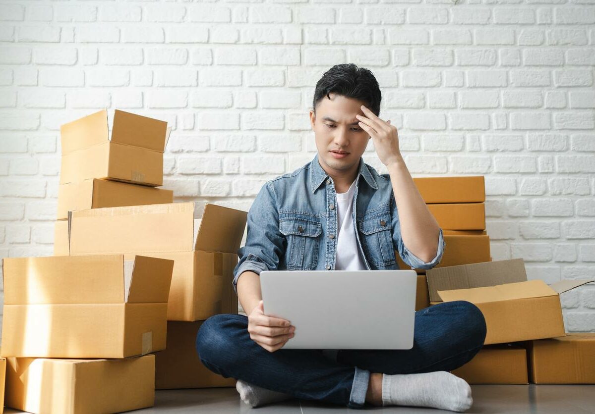 A man who is stressed looks at a laptop while sitting on the floor near boxes
