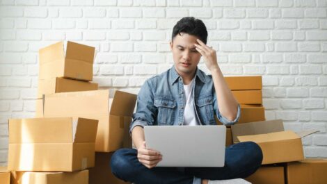 A man who is stressed looks at a laptop while sitting on the floor near boxes