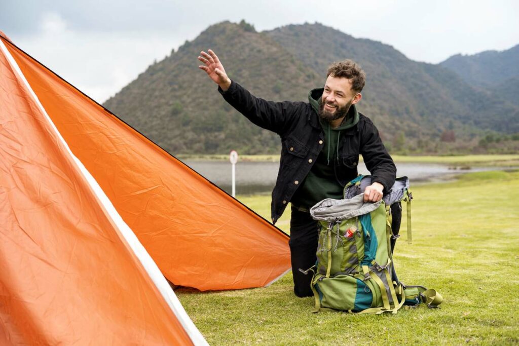 Man waves as he packs his gear next to an orange tent on a grassy field.