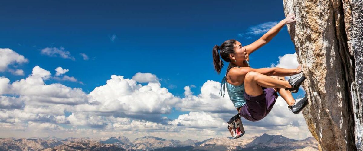 A woman scales the side of a cliff with mountains in the background.