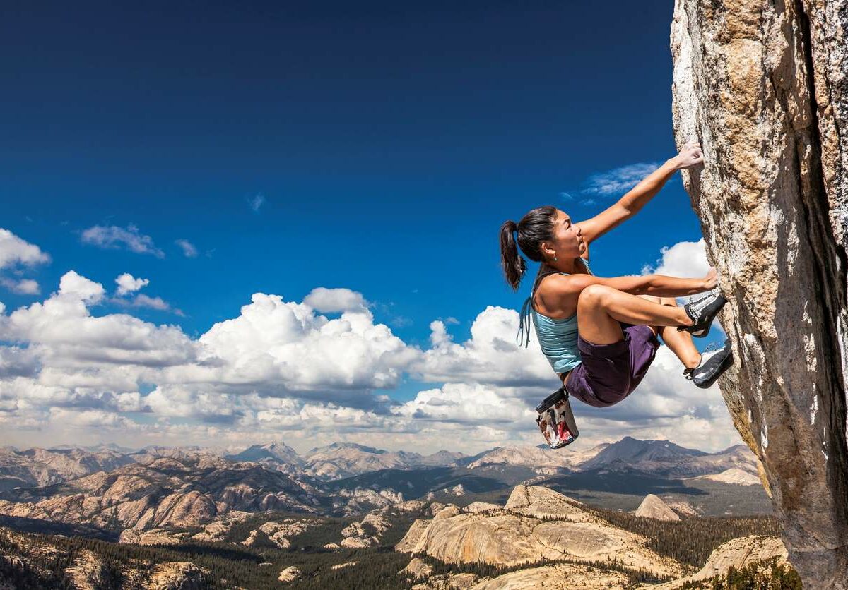 A woman scales the side of a cliff with mountains in the background.