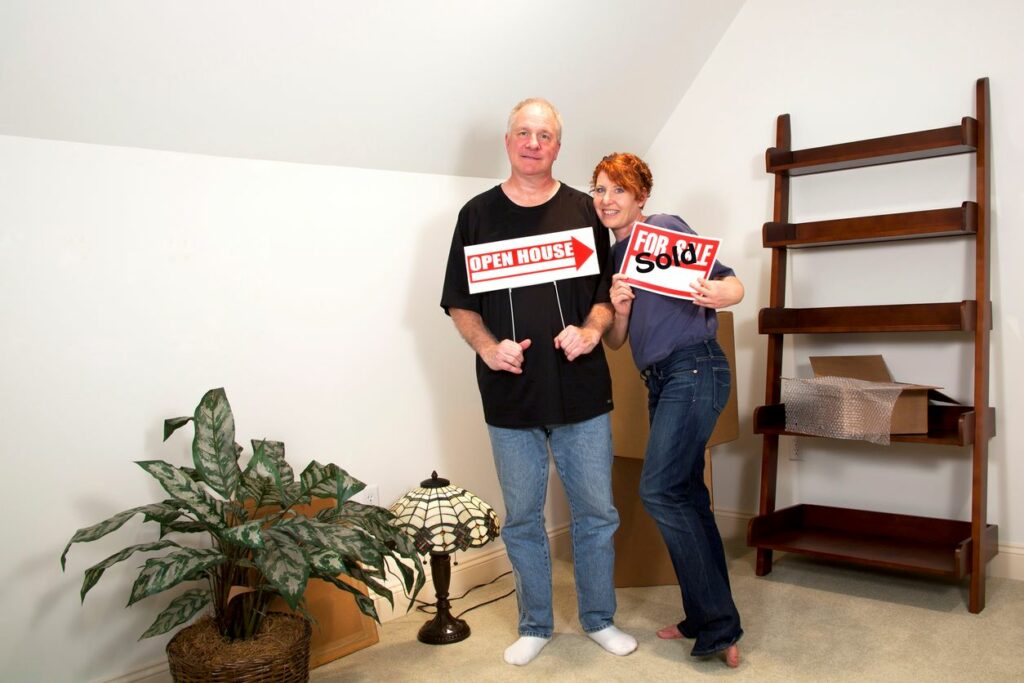 Senior couple standing together inside new home holding “open house” and “sold” signs.