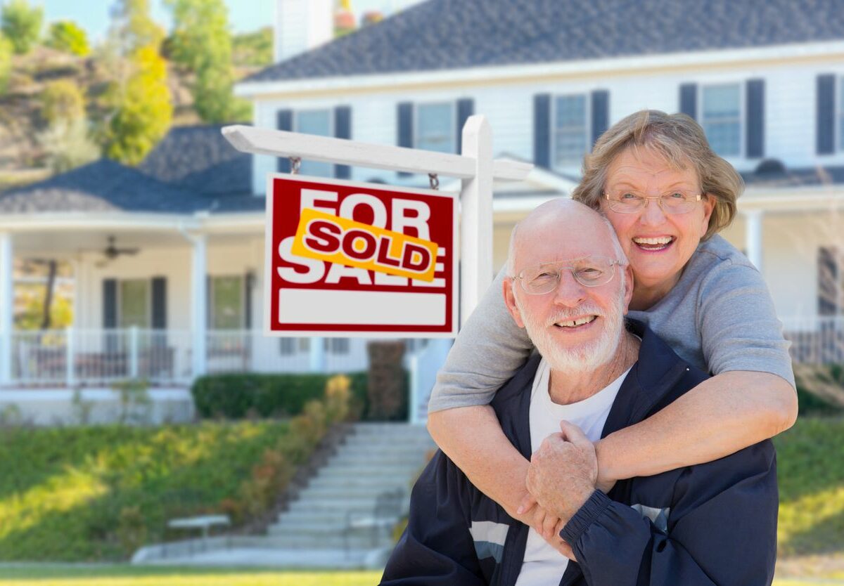 an and woman smiling and hugging beside a sold sign outside their old home as they downsize for retirement.