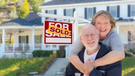 an and woman smiling and hugging beside a sold sign outside their old home as they downsize for retirement.