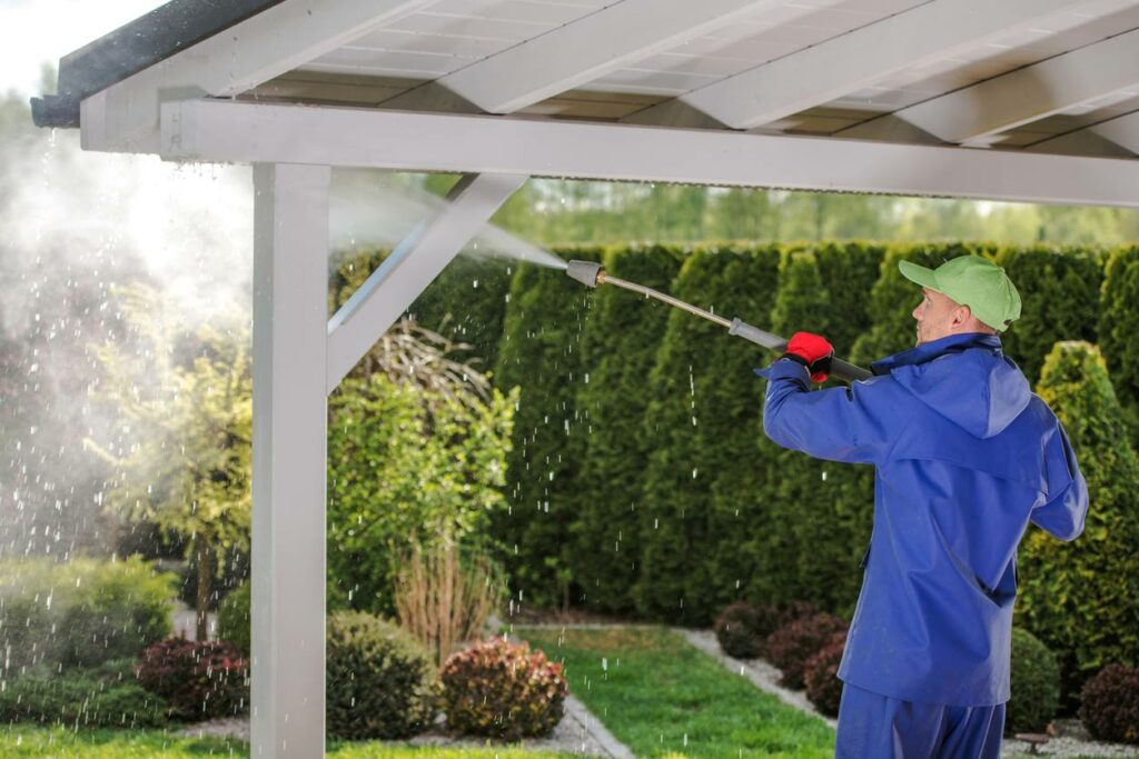 A man pressure washes his porch awning while wearing a protective blue jacket.