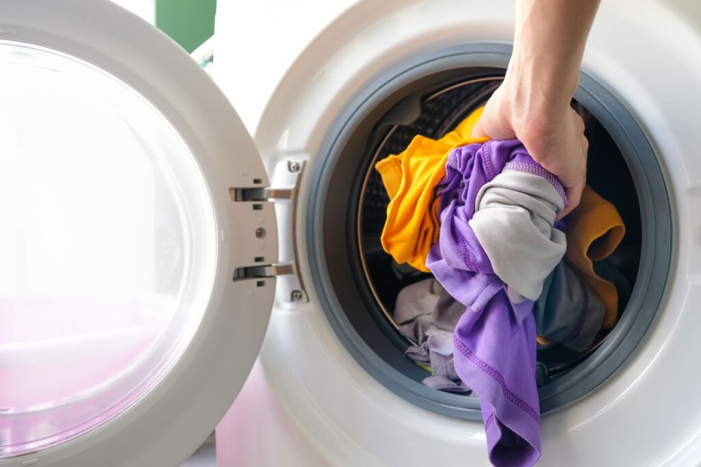 A hand loads clothing into a white washing machine.